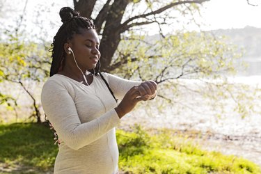 Person in a park wearing a white long sleeve shirt with wired headphones in their ears pausing their watch and taking a break after exercising.